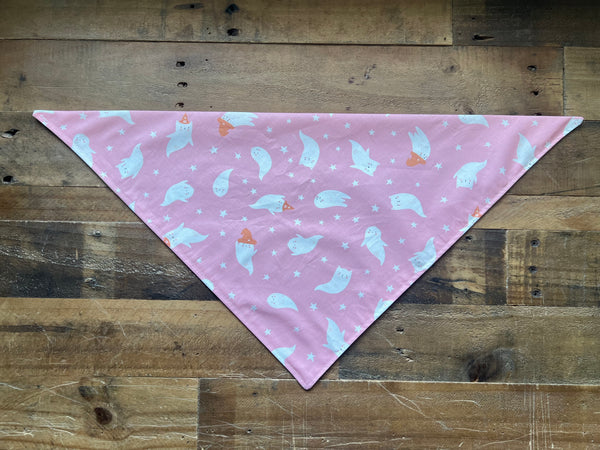 A pink triangular pet bandana with a white ghost and star pattern, laid flat on a rustic wooden plank surface.