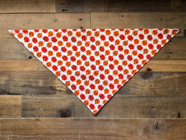 A triangular bandana with a repeating pattern of orange jack-o'-lantern faces on a white background, laid flat on a rustic brown wooden plank surface.