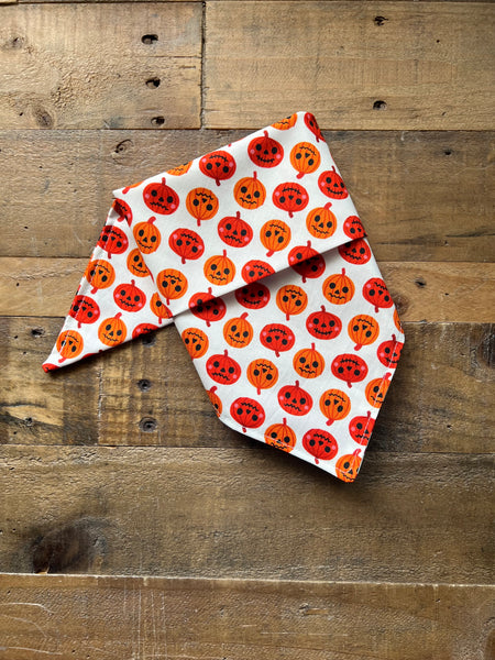 A white bandana-style pet scarf with an all-over orange pumpkin print, laid flat on a rustic wooden plank surface.
