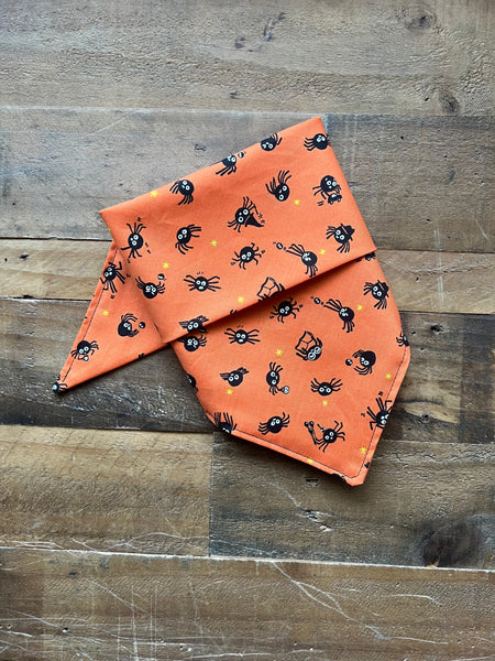 An orange pet bandana with a black spider pattern, laid flat on a rustic wooden surface.