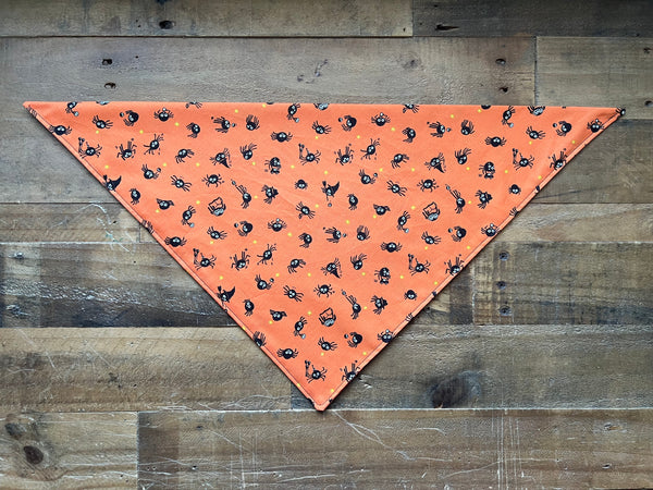 An orange triangular pet bandana with a black spider pattern, laid flat on a rustic wooden plank background.