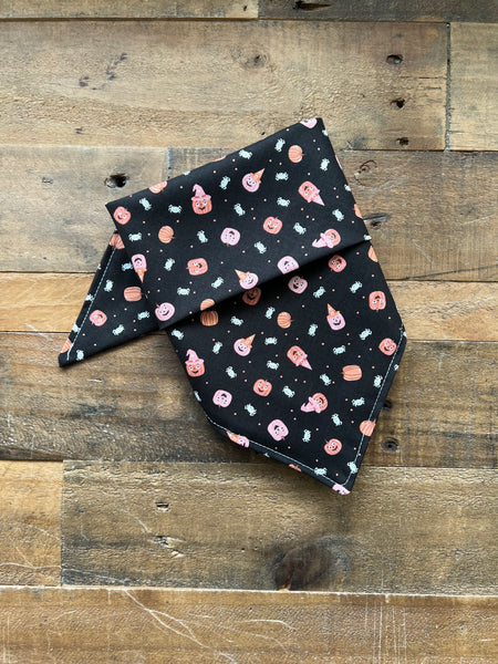 A black pet bandana with a Halloween pattern of orange pumpkins, pink pumpkins, and white spiders, displayed on a rustic wooden plank surface.