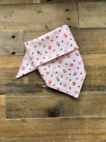 A light pink pet bandana with a cute print of spiders, bats, and pumpkins, folded and displayed on a rustic wooden plank background.