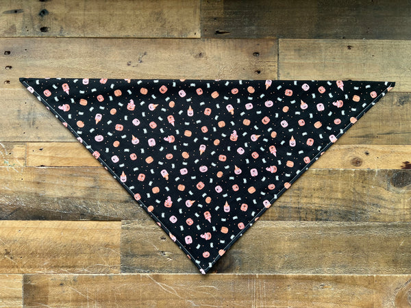 A black triangular pet bandana with a pink, orange, and white pattern, laid flat on a rustic wooden plank background.