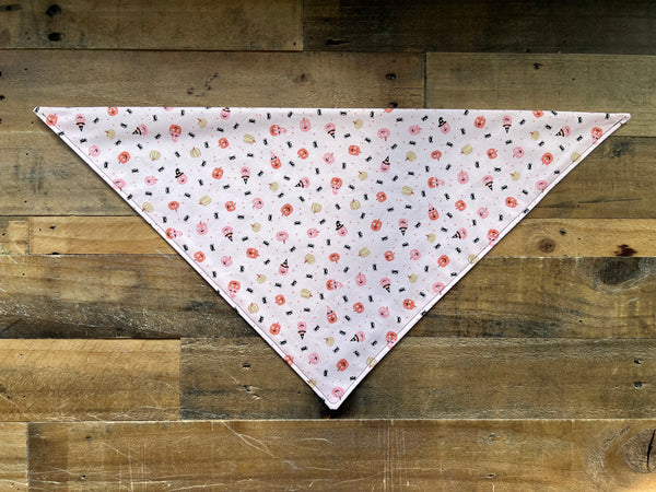 A triangular pet bandana with a pink background and a dense pattern of small Pumpkins, spiders laid flat on a rustic wooden plank surface.