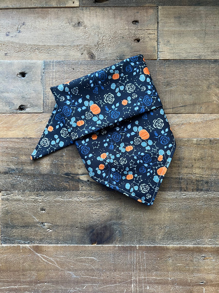 A folded bandana-style pet collar or scarf with a black background and a colorful floral and pumpkin pattern, laid flat on a rustic wooden plank surface.