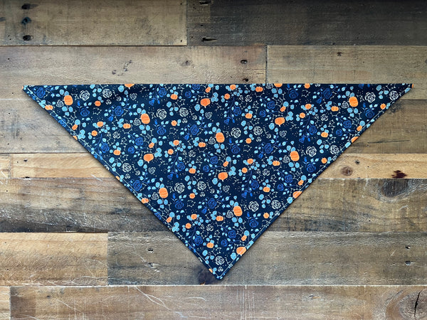 A triangular pet bandana with a black background and a colorful floral and pumpkin pattern, laid flat on a rustic wooden plank surface.