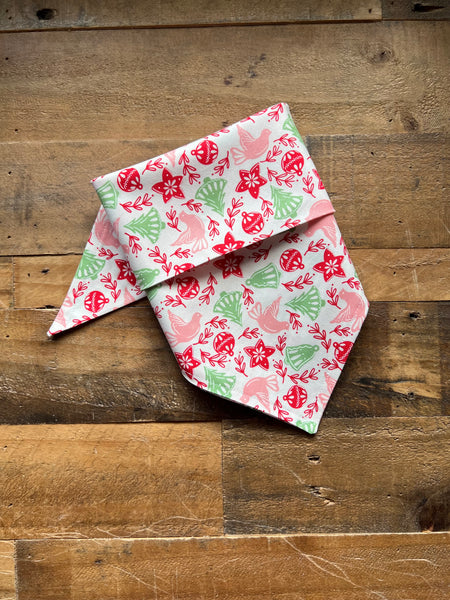 A folded bandana with a festive Christmas pattern featuring red Stars, green pine trees, and pink birds on a white background, displayed on a rustic wooden surface.