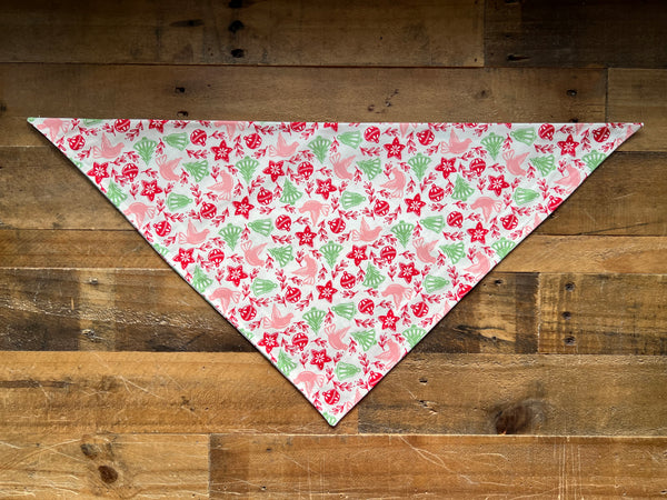 A triangular bandana  with a pink, red, and green holiday pattern, laid flat on a rustic wooden plank background.