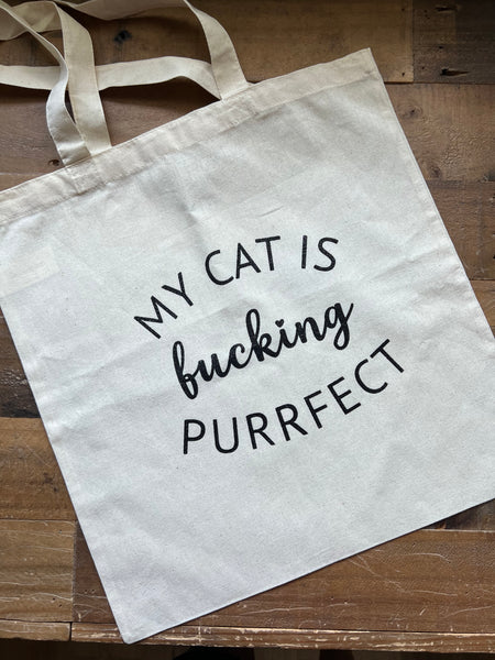 Tote bag with 'My cat is fucking purrfect' text on a wooden surface