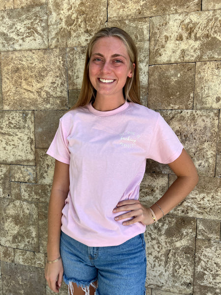 Woman wearing a light pink t-shirt with white text My Dog Is Fucking Perfect and blue jeans standing against a stone wall.