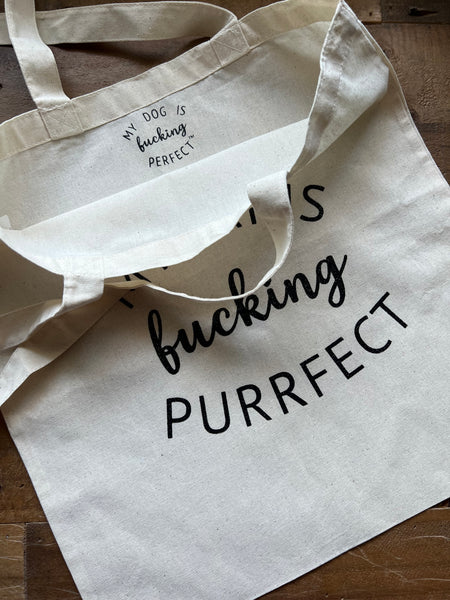 Beige tote bag with My Cat Is Fucking Purrfect on a wooden surface