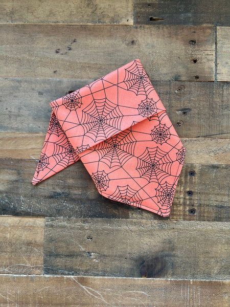 An orange bandana with a black spiderweb pattern, folded and displayed on a rustic wooden plank background.
