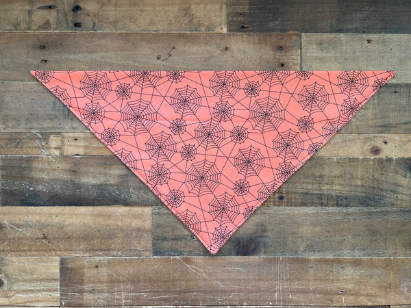 An orange triangular bandana with a black spiderweb pattern, laid flat on a rustic brown wooden plank background.