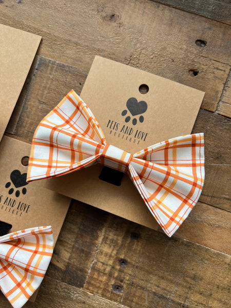 A plaid pet bow tie in orange, white, displayed on a brown kraft paper card with the brand name 'Pets and Love printed on it, set against a rustic wooden background.