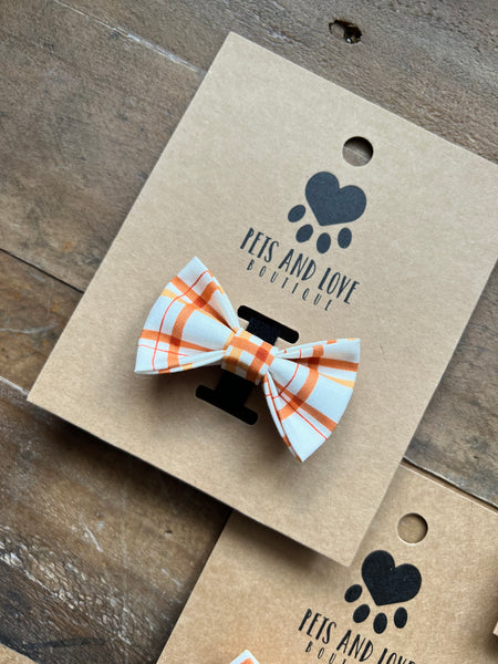 A small pet bow tie with an orange, white, plaid pattern, attached to a brown kraft paper display card printed with the brand name 'PETS AND LOVE BOUTIQUE'. The card is placed on a rustic wooden surface.