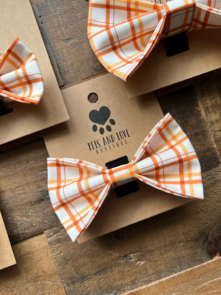 An orange and white plaid pet bow tie displayed on a brown kraft paper tag with a black paw print logo that reads 'KIS AND LOVE PET GIFT STORE'. Multiple similar tags with matching bow ties are arranged on a rustic wooden surface.