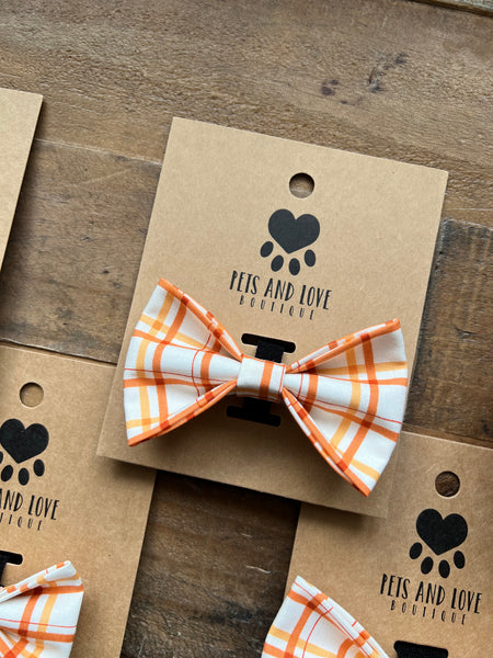An orange and white plaid pet bow tie displayed on a brown kraft paper card with the 'Pets and Love Boutique' logo, arranged on a rustic wooden surface.