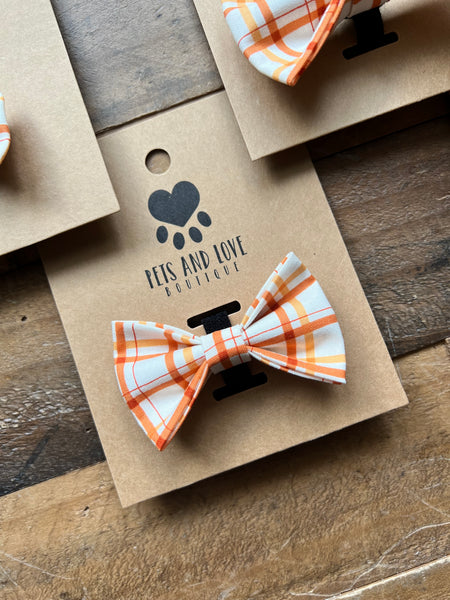 An orange and white plaid dog bow tie displayed on a brown kraft paper card with the brand name 'PETS AND LOVE BOUTIQUE' printed on it, set on a rustic wooden surface.