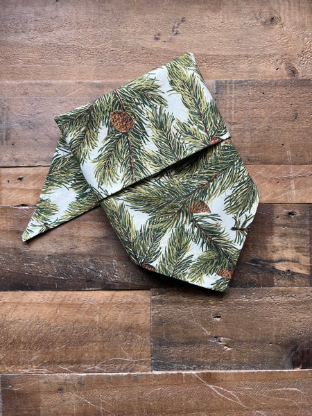 A fabric bandana with a pine branch and pinecone pattern, laid flat on a rustic wooden surface.