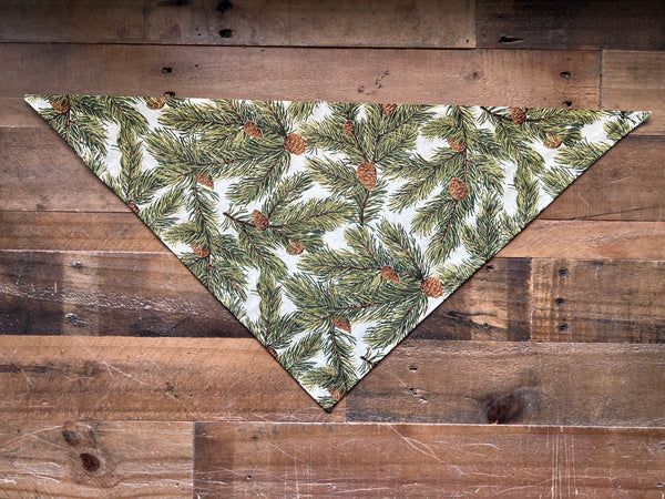 A triangular bandana with a pine branch and pinecone pattern, laid flat on a rustic wooden plank background.