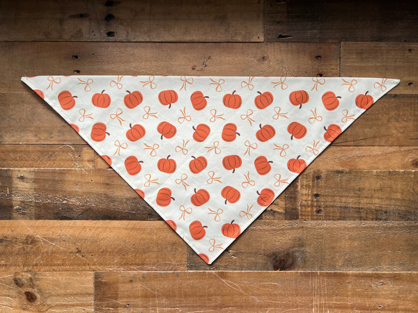 A triangular pet bandana with an orange pumpkin and bow pattern on a beige background, laid flat on a rustic brown wooden surface.