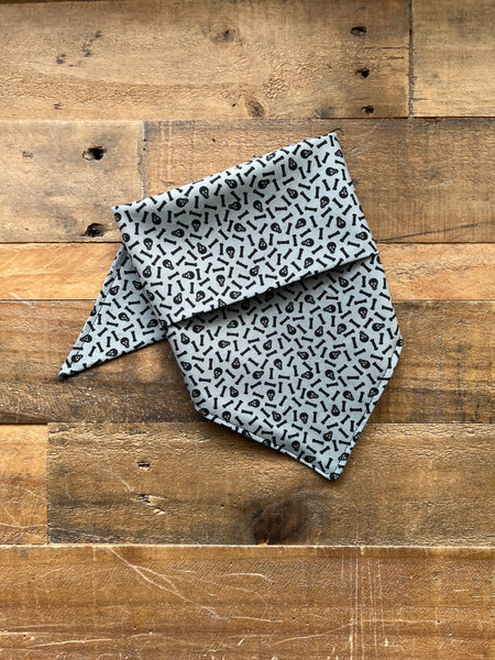 A light gray bandana with Tiny black skulls and bones pattern, laid flat on a rustic wooden plank background.