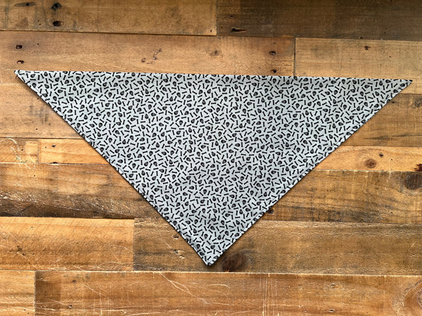 A triangular bandana gray background with tiny black skulls and bones, laid flat on a rustic wooden plank background. 