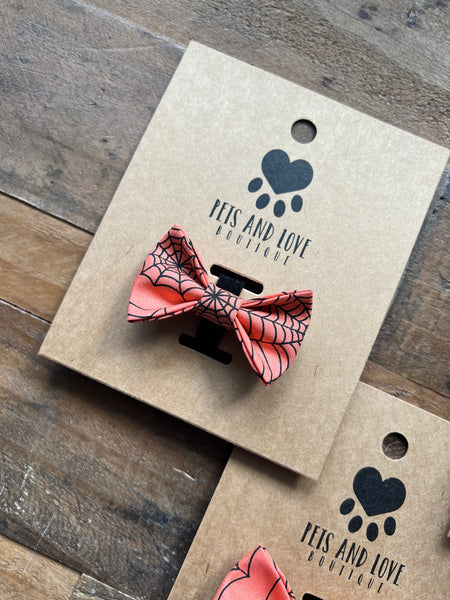 A orange and black spiderweb-patterned pet bow tie displayed on a brown kraft paper card with the brand name 'PETS AND LOVE BOUTIQUE' printed on it, set on a rustic wooden surface.