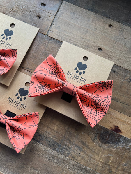 Orange pet bowties with a black spiderweb pattern, displayed on brown kraft paper cards with the brand name 'Pets and Love Boutique' printed on them, all arranged on a rustic wooden surface.