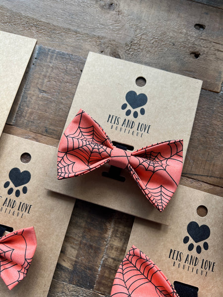 An orange and black spiderweb-patterned bow tie displayed on kraft paper cards printed with the brand name 'PETS AND LOVE BOUTIQUE', arranged on a rustic wooden surface.