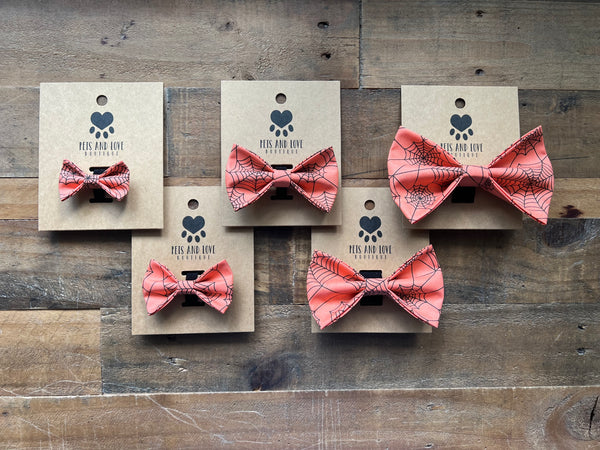 Five pet bow ties with an orange and black spiderweb pattern, each mounted on a kraft paper display card with a paw print logo, arranged on a rustic wooden surface.