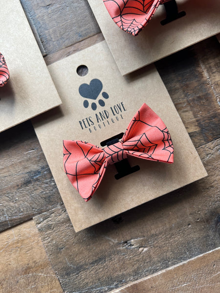 An orange and black spiderweb-patterned bow tie displayed on a brown kraft paper card with the brand name 'PEIS AND LOVE BOUTIQUE' printed on it, set on a rustic wooden surface.