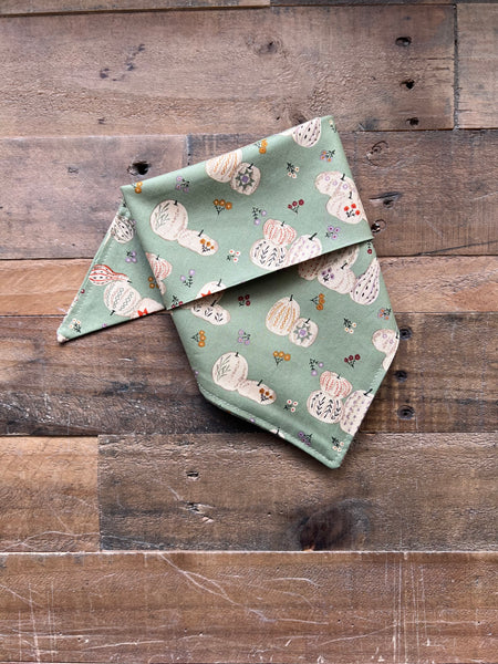 A sage green bandana-style pet collar with a pumpkin and flower pattern, laid flat on a rustic wooden plank background.