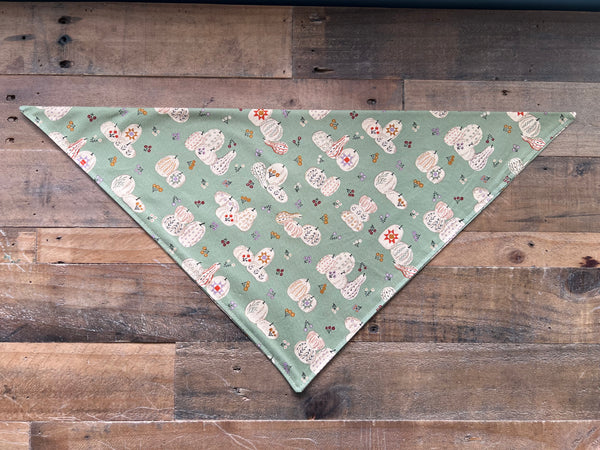 A triangular pet bandana with a sage green background and a repeating pattern of beige pumpkins and small orange and purple flowers, laid flat on a rustic wooden plank surface.
