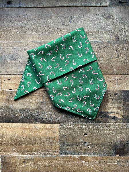 A green fabric pet bandana with a repeating candy cane pattern, folded and displayed on a rustic wooden plank surface.