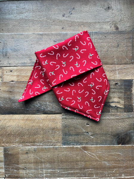 A red bandana with a repeating white and green candy cane and holly pattern, folded and displayed on a rustic wooden plank surface.