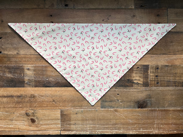 A triangular bandana laid flat on a rustic wooden plank surface, featuring a cream background with a repeating red and green candy cane print.