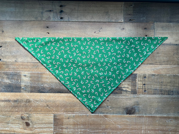 A green triangular pet bandana with a white candy cane pattern, laid flat on a rustic wooden plank background.