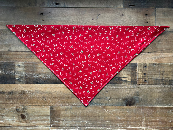 A red triangular pet bandana with a white and green candy cane pattern, laid flat on a rustic wooden plank background.