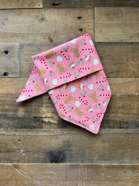 A pink pet bandana with a holiday gingerbread print, laid flat on a rustic wooden plank background.