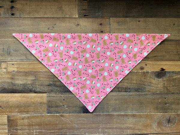 A pink triangular bandana with a repeating holiday cookies Pattern, laid flat on a rustic wooden plank background.