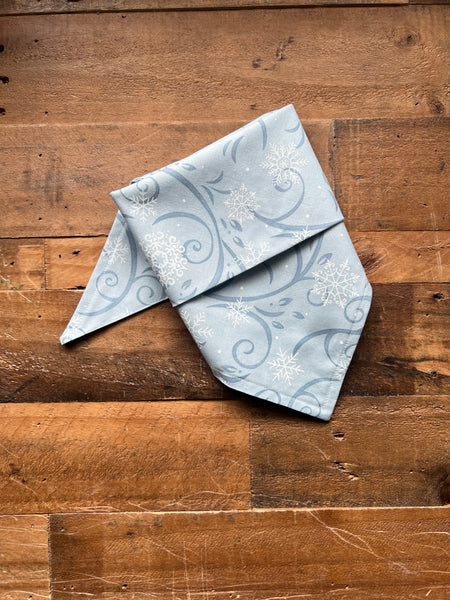 A light blue bandana with a white snowflake and swirl pattern, folded and displayed on a rustic wooden surface.