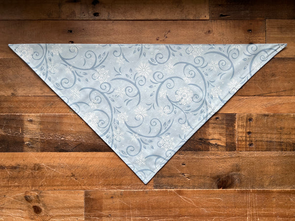 A triangular bandana with a light blue and white snowflake pattern, laid flat on a rustic brown wooden plank background.