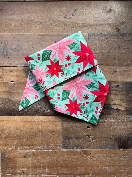 A folded fabric pet bandana with a festive holiday print of red poinsettias, pink flowers, and green leaves on a mint green background, displayed on a rustic wooden plank surface.