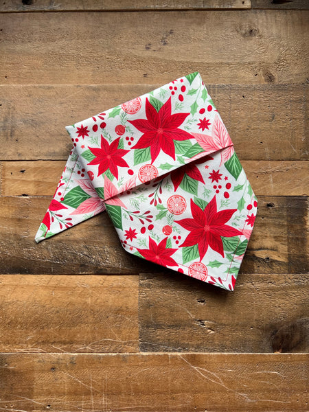 A folded white fabric pet bandana with a red pink and green Christmas poinsettia and berry pattern, displayed on a rustic wooden plank surface.
