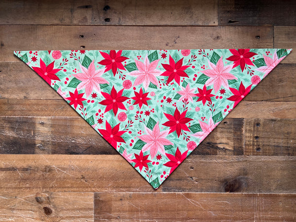 A triangular bandana with a festive Christmas poinsettia pattern, laid flat on a rustic wooden plank background.