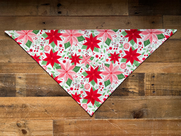 A triangular pet bandana with a festive holiday print of red poinsettias, pink flowers, and green leaves on a white background, laid flat on a rustic wooden plank surface.